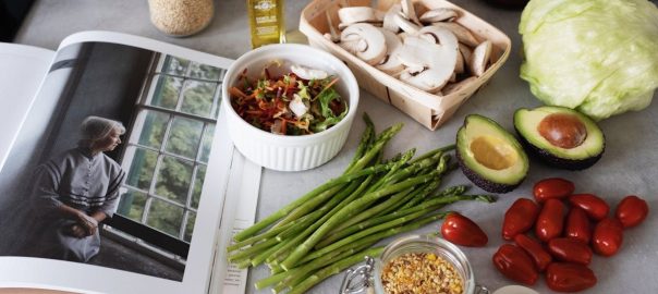 A selection of fresh vegetables and a cookbook in a kitchen setting, promoting healthy cooking.